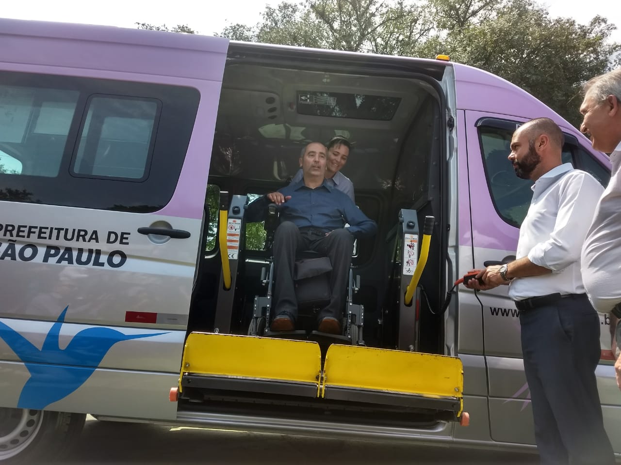 Uma mulher auxiliando uma pessoa em cadeira de rodas sendo retirada da van do serviço Atende+, do lado de fora dois homens, um operando o elevador da van e outro observando a ação.